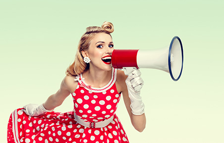 Woman holding megaphone, dressed in pin-up style red dress in polka dot, on green background. Caucasian blond model posing in retro fashion vintage studio shoot. Copyspace area for advertising slogan or text message.
